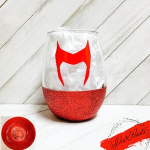 Op de afbeelding: Rood glitter wijnglas met een rood Scarlet Witch-logo. Het glas is helder met een rode glitterbodem.