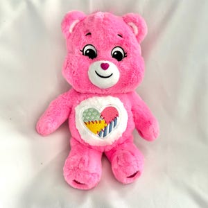 May include: Pink plush Care Bear with a patchwork heart on its chest. The heart is made of different colored fabric pieces and has stitching around the edges.