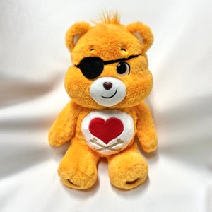 Könnte beinhalten: Ein gelbes Care Bear Plüschtier mit einem schwarzen Augenklappen, einem roten Herzen auf der Brust und gekreuzten Knochen unter dem Herzen. Der Care Bear lächelt.