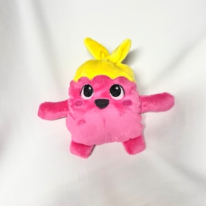 Puede incluir: Juguete de peluche rosa con una diadema amarilla. El juguete tiene una cara sonriente con ojos negros y una boca negra.