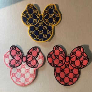 May include: Three embroidered patches in the shape of a mouse head with a bow. The top patch is navy blue with gold accents. The other two are pink and red with black accents. The patches feature a repeating logo pattern.