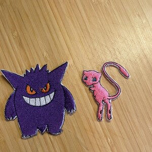 May include: Two embroidered patches featuring cartoon characters. One is a purple creature with a wide grin and orange eyes. The other is a pink cat-like creature with a long tail.
