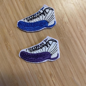 May include: Two embroidered patches shaped like sneakers. One patch has a white upper with black details and a blue sole. The other patch has a white upper with black details and a purple sole. Both patches are on a wooden surface.