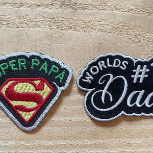 May include: Two embroidered fabric patches. The left patch is shield-shaped with a black background, green text reading SUPER PAPA, and a red and yellow S emblem. The right patch is black with white text reading WORLDS #1 Dad in script.