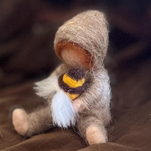 Könnte beinhalten: Eine handgefertigte Filzpuppe, die ein Kapuzenoutfit in Braun- und Beigetönen trägt. Die Puppe hält ein gelbes, braunes und weißes Filzobjekt. Die Puppe sitzt auf einer braunen Oberfläche.