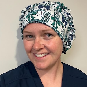 May include: A white scrub cap featuring a repeating pattern of dark blue and teal bows, baseballs, and the text Mariners in a stylized font. The cap has a gathered, elasticized back design and is constructed from a lightweight fabric.