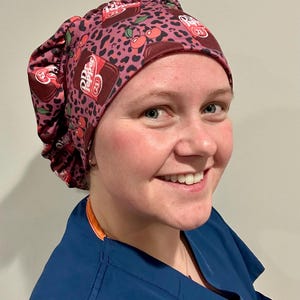May include: A burgundy scrub cap with a Dr. Pepper and cherry print. The cap features a leopard print background and the Dr. Pepper logo with the number 23. The cap is worn by a person wearing a blue scrub top.