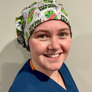 Puede incluir: Un gorro quirúrgico estampado en blanco y gris con el texto "MTN DEW AND JESUS" y logotipos de Mountain Dew y cruces. Lo lleva una persona con una bata quirúrgica azul.
