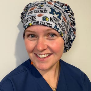 Puede incluir: Un gorro quirúrgico con fondo gris claro y un patrón repetitivo de logotipos de Michigan Wolverines, el texto "Michigan" y "Wolverines" y balones de fútbol americano marrones. El gorro tiene un diseño redondeado y fruncido con una banda ancha.