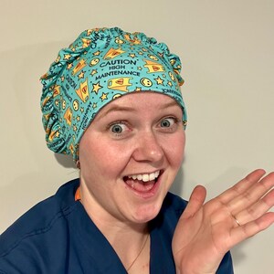 Può includere: Un cappello chirurgico turchese con un motivo ripetuto di segnali di pericolo gialli, stelle e faccine sorridenti. Il testo "CAUTION HIGH MAINTENANCE" è stampato sul cappello. Il cappello è indossato da una persona.