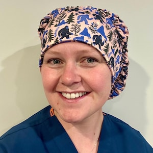 May include: A scrub cap featuring a light pink background with a repeating pattern of dark blue Bigfoot silhouettes, green pine trees, and blue mountain shapes. The cap has a gathered, elasticized back design to provide a secure and adjustable fit.