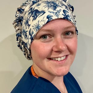 May include: A white and blue patterned surgical scrub cap featuring a dinosaur toile design. The cap has a ruffled edge and is made of a soft fabric. The cap is worn by a person wearing a blue scrub top.