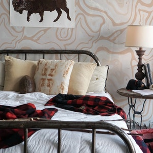 May include: A white bed with a black metal frame and a white comforter. There are red and black plaid blankets on the bed. There are pillows on the bed, including a brown and white pillow with a pattern. The bed is in front of a wall with a brown and white abstract pattern. There is a brown and white picture of a buffalo on the wall. There is a lamp on a table next to the bed.