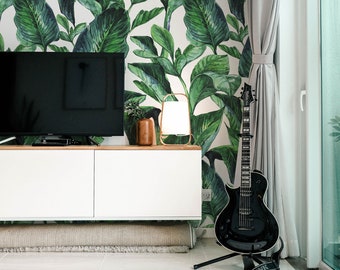 May include: A living room interior with a white wall covered in a green tropical leaf wallpaper. A black electric guitar is leaning against a white curtain. A white entertainment center with a flat screen TV sits below the wallpaper. A brown wooden coffee table is in the foreground.