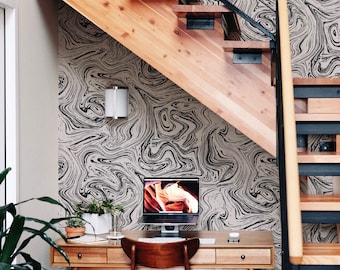 Op de afbeelding: Een thuiskantoorwerkplek met een houten bureau en stoel, een computermonitor en planten. De muur achter het bureau is bedekt met zwart-wit marmerbehang. Een houten trap met zwarte metalen leuningen bevindt zich op de achtergrond.