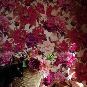 K&ouml;nnte beinhalten: Eine schwarze Katze sitzt neben einem geflochtenen Korb, der mit rosa und roten Blumen gef&uuml;llt ist. Der Korb steht vor einer Wand mit einem floralen Tapetenmuster. Eine braune Strickdecke liegt auf dem Boden.