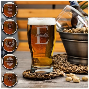 May include: A personalised beer glass with the name "EDWARD" etched on the side. The glass is filled with a golden-coloured beer and is sitting on a woven coaster. There are pistachios scattered on a wooden surface.