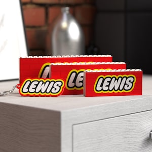 May include: Red Lego-style brick-shaped items with the name "LEWIS" in white letters on a yellow background. The items vary in size, with one attached to a keychain. The items are displayed on a white surface.