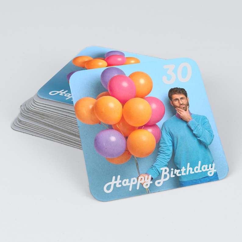 Peut inclure: Une pile de dessous de verre carr&eacute;s sur fond bleu clair, montrant une personne en pull bleu tenant un bouquet de ballons color&eacute;s. Le texte 30 et Happy Birthday est imprim&eacute; en blanc. Les dessous de verre ont des coins arrondis et une bordure.