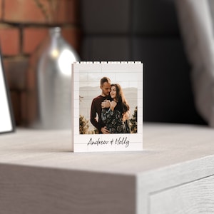 May include: A white Lego-style photo frame with a picture of a couple embracing. The frame has a white background with black brick outlines and the text "Andrew & Holly" written below the photo.