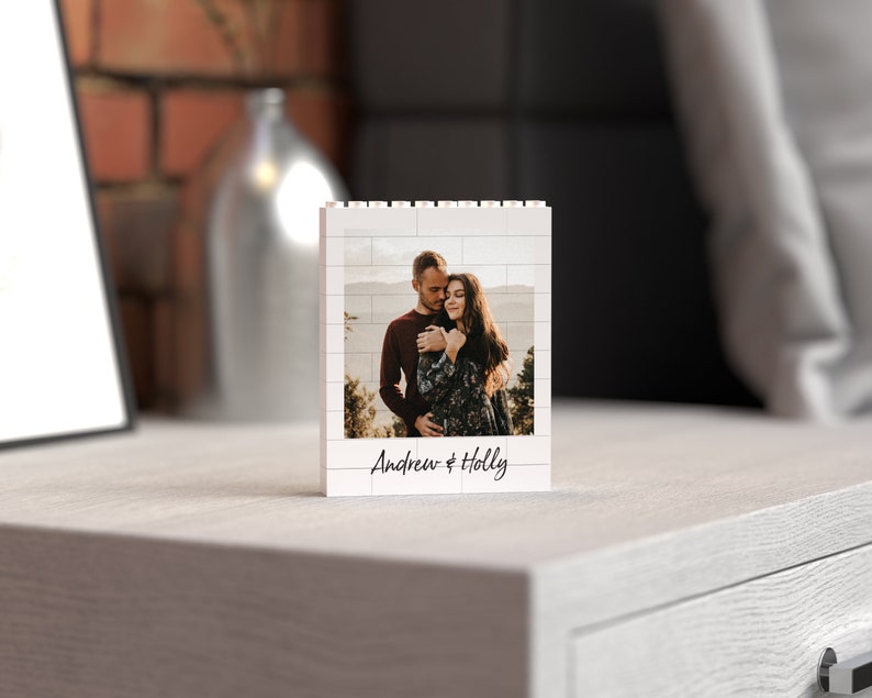 May include: A white Lego-style photo frame with a picture of a couple embracing. The frame has a white background with black brick outlines and the text "Andrew & Holly" written below the photo.