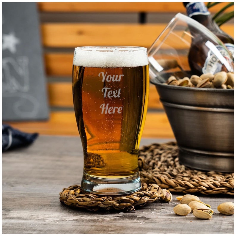 May include: A clear glass beer mug with a frosted finish, filled with a golden beer. The mug is on a woven coaster and has the text "Your Text Here" etched on the side.