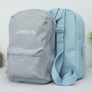 May include: Two backpacks, one grey and one light blue. The grey backpack has the name "James." embroidered on the front.
