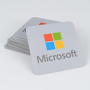 Peut inclure: Une pile de dessous de verre carr&eacute;s gris clair arborant le logo Microsoft en rouge, vert, bleu et jaune, avec le mot Microsoft imprim&eacute; en gris sous l'ic&ocirc;ne. Les dessous de verre ont des coins arrondis et une surface mate et plate.