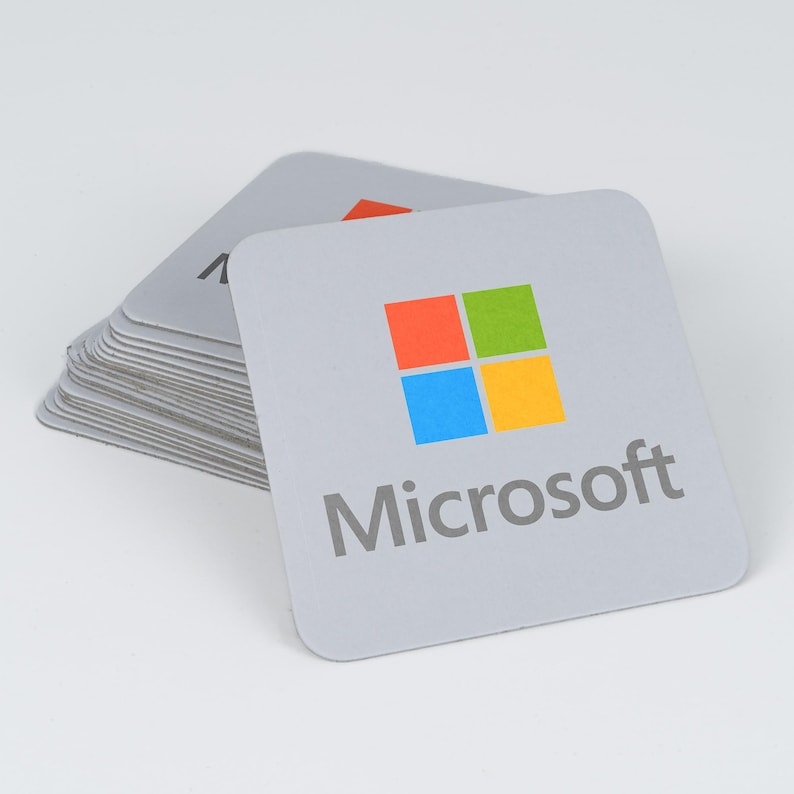 Peut inclure: Une pile de dessous de verre carr&eacute;s gris clair arborant le logo Microsoft en rouge, vert, bleu et jaune, avec le mot Microsoft imprim&eacute; en gris sous l'ic&ocirc;ne. Les dessous de verre ont des coins arrondis et une surface mate et plate.