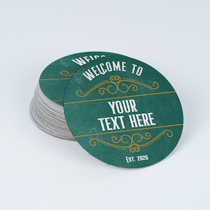 Peut inclure: Une pile de sous-verres circulaires sur fond bleu sarcelle fonc&eacute;. Le sous-verre sup&eacute;rieur pr&eacute;sente le texte blanc "WELCOME TO", "YOUR TEXT HERE" et "EST. 2026". Des fioritures d&eacute;coratives dor&eacute;es et des lignes horizontales encadrent le texte.