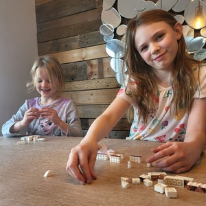 Op de afbeelding: Twee jonge meisjes zitten aan een houten tafel en bouwen met witte en bruine speelgoedblokken. Een meisje glimlacht terwijl ze aan haar creatie werkt. De achtergrond toont een houten muur en een decoratieve spiegel.
