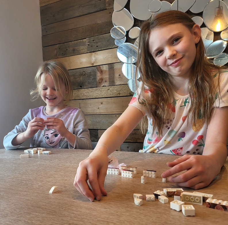Op de afbeelding: Twee jonge meisjes zitten aan een houten tafel en bouwen met witte en bruine speelgoedblokken. Een meisje glimlacht terwijl ze aan haar creatie werkt. De achtergrond toont een houten muur en een decoratieve spiegel.