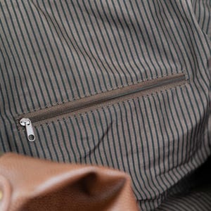 May include: Close-up of a brown leather bag with a zippered pocket. The pocket is lined with a brown and black striped fabric.