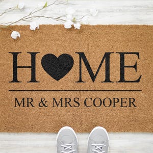 May include: A brown coir doormat with the word "HOME" in black lettering with a black heart in place of the "O". The text "MR & MRS COOPER" is printed below the word "HOME".
