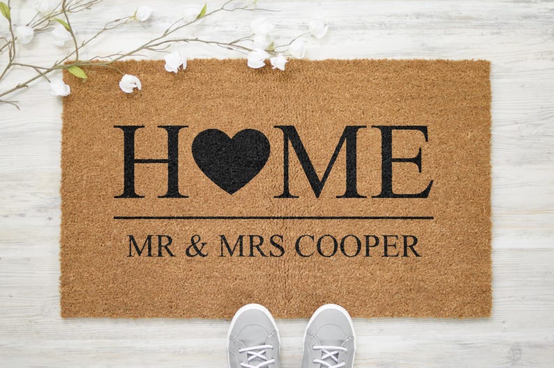May include: A brown coir doormat with the word "HOME" in black lettering with a black heart in place of the "O". The text "MR & MRS COOPER" is printed below the word "HOME".