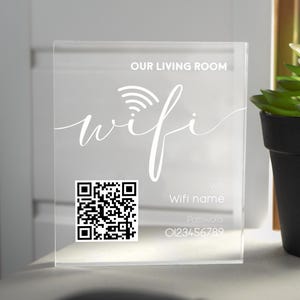 May include: A clear acrylic sign with the words "Our Living Room Wifi" and a wifi symbol. A black and white QR code is below the text, along with the words "Wifi name" and "Password" and a number.