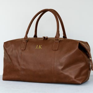 May include: A brown leather duffel bag with a gold zipper and gold initials "J.K." embroidered on the front.