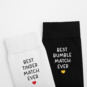 May include: A pair of socks with a white sock that says "Best Tinder Match Ever" with a red heart and a black sock that says "Best Bumble Match Ever" with a yellow heart.