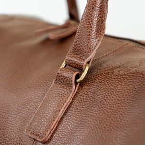 May include: Close-up of a brown leather duffel bag with a gold metal buckle. The bag has a textured surface and a top handle.