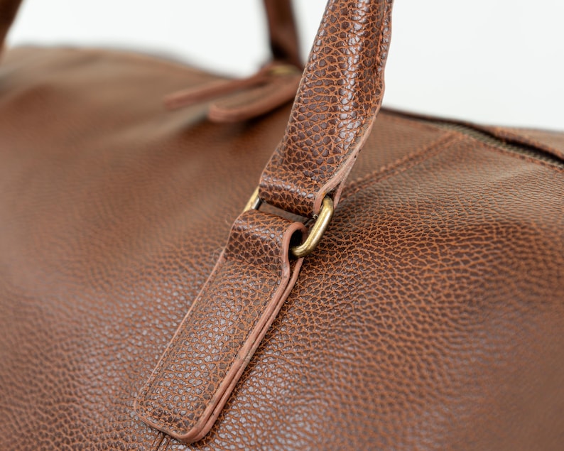 May include: Close-up of a brown leather duffel bag with a gold metal buckle. The bag has a textured surface and a top handle.