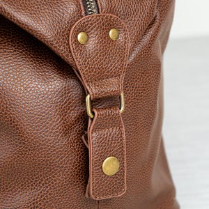May include: Close-up of a brown leather duffel bag with a brass zipper and buckle. The bag has a textured surface and a flap with two brass studs.