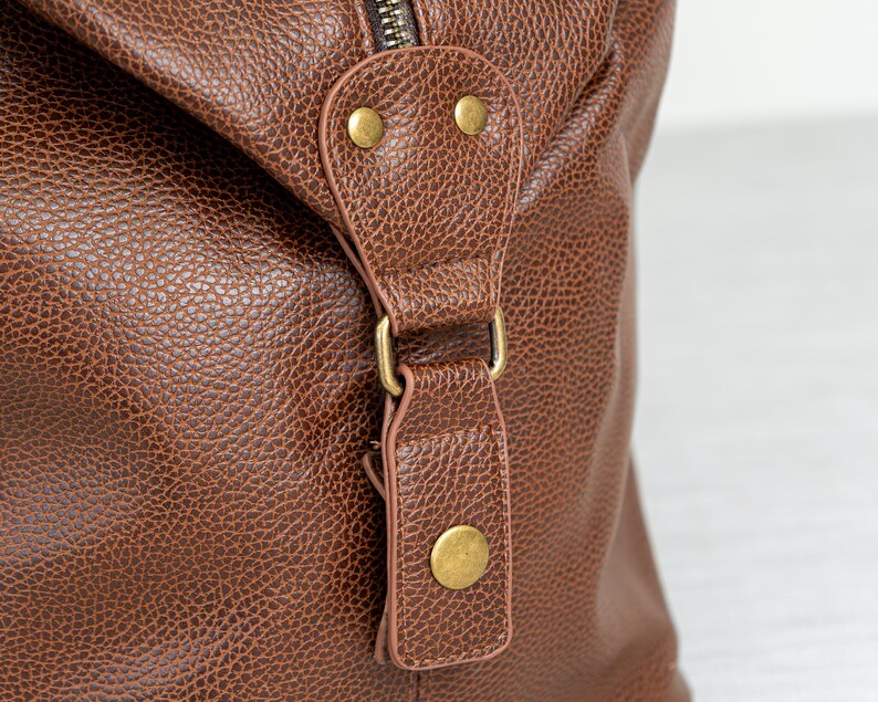 May include: Close-up of a brown leather duffel bag with a brass zipper and buckle. The bag has a textured surface and a flap with two brass studs.