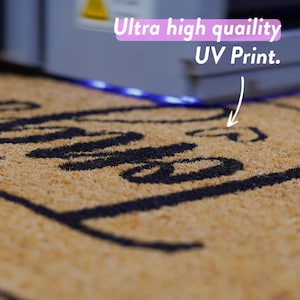 Puede incluir: Primer plano de una alfombra de coco marr&oacute;n con letras negras que dicen "Hello". La alfombra est&aacute; iluminada por una tira de luz LED azul. El texto "Ultra high quality UV Print." es visible en el fondo.