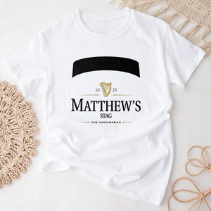 May include: White t-shirt with a black and gold design. The design includes the text "MATTHEW'S STAG" with "THE GROOMSMAN" below. A black banner and a gold harp are also featured. The shirt is laid flat on a white surface.