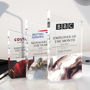 May include: Three clear acrylic awards with printed text and abstract designs. The awards are for "Barista of the Year," "Manager of the Year," and "Employee of the Month." Each award features a different company logo and recipient name.