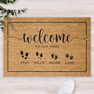 Puede incluir: Felpudo rectangular de fibra de coco natural. El felpudo presenta las palabras "welcome to our home" en escritura negra, junto con huellas de pies y patas. Los nombres "Ryan", "Millie", "Archie" y "Luna" están impresos debajo.