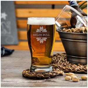 May include: A personalized beer glass with the name "Helen Bull" etched on it. The glass is filled with a golden-colored beer and has a floral design etched on the front.