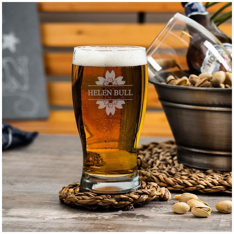 May include: A personalized beer glass with the name "Helen Bull" etched on it. The glass is filled with a golden-colored beer and has a floral design etched on the front.