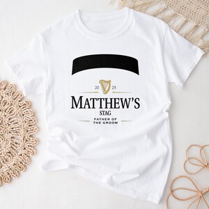May include: White t-shirt with a black and gold design. The design includes the name "Matthew's", the word "Stag", and "Father of the Groom". The design also features a black banner and a gold harp symbol.