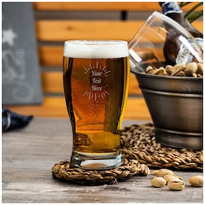 May include: A clear glass beer mug with a frosted design that says "Your Text Here". The mug is filled with a golden beer and is sitting on a woven coaster.